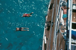 A two people dive into clear sea while a man sunbathes on the deck of a Swan yacht