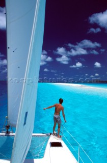 Man stands look out on the bow of a cruising catamaran approaching blue shallow water and a sandy spit on Pine Island, New Caledonia