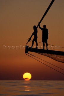 Couple standing on bowsprit of classic yacht at sunset.