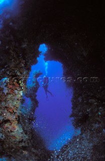 Scuba diver ascends from an underwater cave surrounded by coral, expelling air bubbles in the blue water as he rises