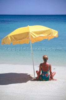 Woman sits alone on beach lookin out to sea under the shade of a yellow umbrella