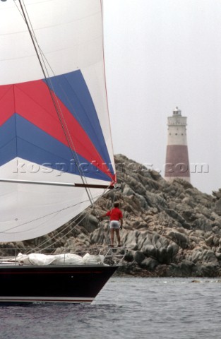 Bowman on foredeck of sailing yacht with lighthouse and rocks in background Sardinia