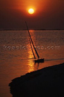 Model yacht sailing off beach at sunset.