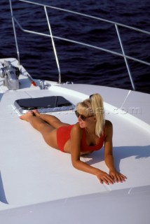 Beautiful blond female girl model sunbathing onboard the bow of a motoryacht in a red bikini