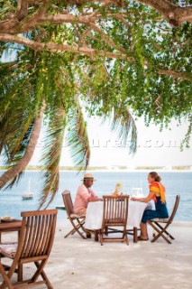 A man and woman sit under shade trees at a table on a patio overlooking the blue water of Lamu Channel.