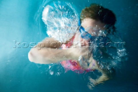 A young girl wearing goggles holding her nose coming up for air amidst her bubbles while swimming in