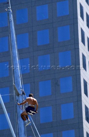 Crew member climbing mast of maxi yacht in front of office block