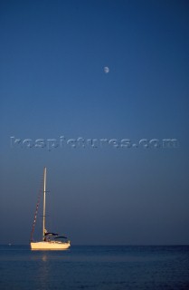 Yacht moored on calm water at sunset with moon in blue sky