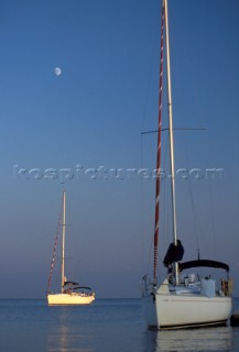 Two yachts moored on calm water at sunset with moon in blue sky