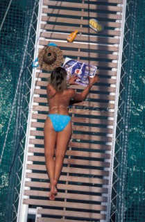 Beautiful suntanned female girl model lying topless on a slated bow sprit reading a magazine in a stylish turquoise blue bikini onboard a traditional boat, with pineapple food and bottle of suncream