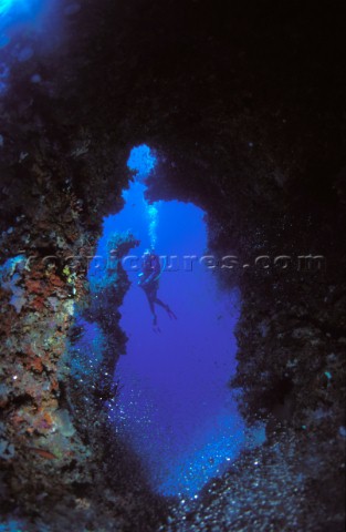 Scuba diver ascends from an underwater cave surrounded by coral expelling air bubbles in the blue wa