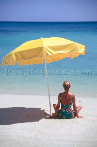 Woman sits alone on beach lookin out to sea under the shade of a yellow umbrella