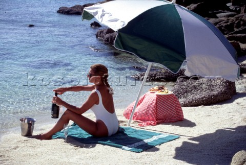 Woman sitting on towel by waters edge opening a bottle of champagne