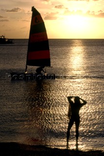 Woman standing on beach watching man in sailing dinghy at sunset