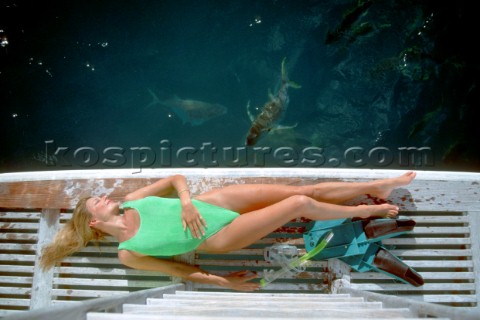Woman lying on dive platform on stern of power boat next to two big fish near the surface of calm wa