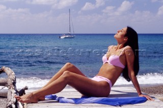 Woman sunbathing on sandy beach with anchored sailing boat in background.