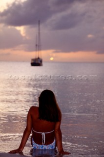 Woman in bikini looking out to sea with anchored sailing yacht in distance