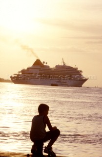 Boy on shore loos out to sea at sunset - cruise ship in background