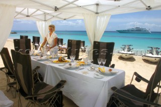 Table layed for lunch under awning on sandy beach with superyacht anchored in distance
