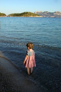 Little girl in pink dress paddling on sandy beach in bay of Formentor, Mallorca