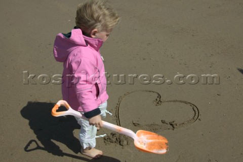 Little girl in pink jacket with bucket and spade drawing heart shape in sand