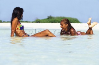 Two girls in bikinis lying in shallow water on sandy beach.