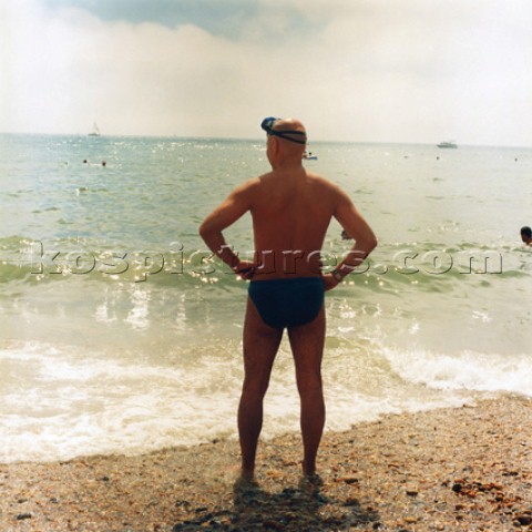 Man in swimming trunks standing on Brighton beach looking out to sea