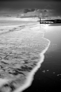 Black and white image of sea lapping onto a hard sand beach with groyne in distance