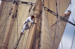 A brave white suited crew man with knife climbs aloft into the masts, sails and rigging of the classic three masted gaff rigged top sail schooner superyacht Adix, to do maintenance and repairs to the fitting on the end of the spinnaker pole below the stainless steel spreaders