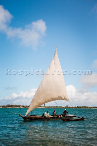 Three men sail a small dhow sailboat in turquoise waters of Lamu Channel with beach in distance