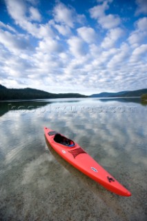 A red sea kayak floats in the shallow reflective waters of Priest Lake in Northern Idaho.  The image was taken in the morning and clouds are reflected on the surface of the lake