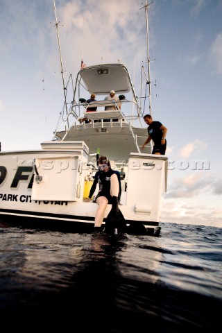 A woman wearing goggles snorkel and flippers sits on the edge of a boats dive door preparing for a d