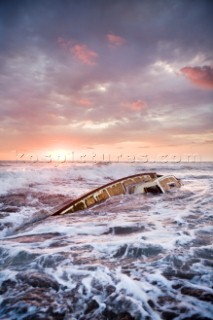 Waves crash over the remains of a sailboat with a dramatic sunset in the background.  The sailboat, the Culin, washed ashore after its owner John Long was murdered by pirates at sea.