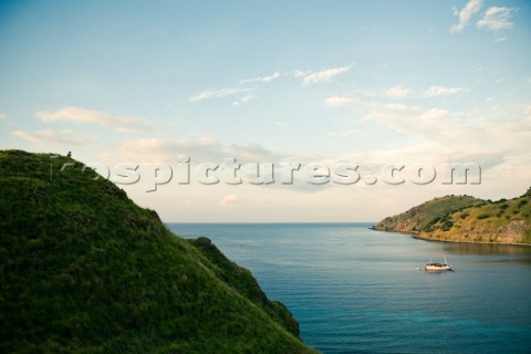 A man sits on the lush green grass covered hillside overlooking his boat in the bay of a small islan