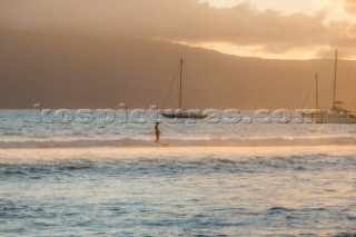 A solo surfer rides a long slow wave with sailboats and an island in the distance off the coast of Maui.
