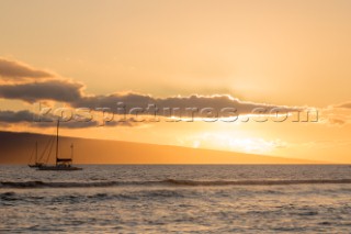 A sailboat at sunset with an island in the distance off the coast of Maui.