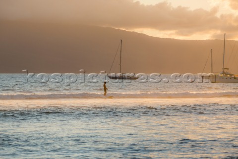 A solo surfer rides a long slow wave with sailboats and an island in the distance off the coast of M