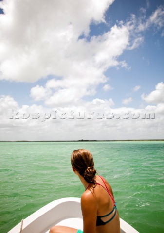 A young woman in a bikini sits in the bow of a white boat as it moves through tealgreen water of the